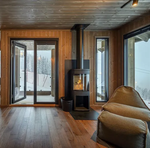 Gemütlicher Holzraum mit schwarzem Kaminofen, Feuer im Sichtfenster, Glastür zum Balkon und Schnee draußen..