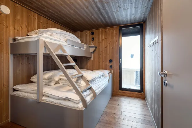 Schmales Holzschlafzimmer mit grauem Etagenbett, Leiter, gestapelten Bettdecken und hohem Fenster zur Terrasse..