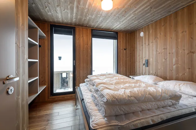 Kleines Schlafzimmer mit Holzverkleidung, Doppelbett mit weißer Bettwäsche und hoher Matratze sowie Tür zum Balkon..