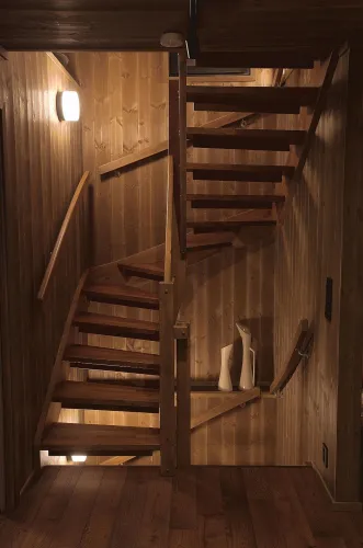 Innenaufnahme eines holzvertäfelten Treppenhauses mit gewendeter Holztreppe, Geländern und warmen Wandlampen..