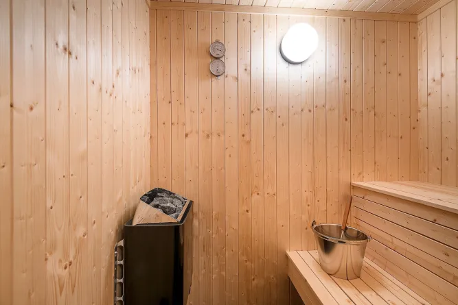 Innenraum einer Holzsauna mit Saunaofen und Steinen, Thermometer-Hygrometer, Bank und Metallkübel mit Kelle..