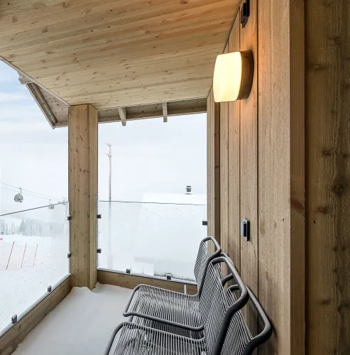 Überdachter Holzbalkon mit zwei Metallstühlen, Glasgeländer und Schnee; im Hintergrund winterliche Skipiste und Gondeln..