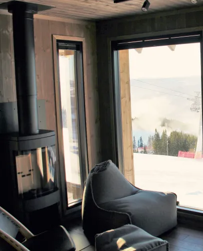 Gemütlicher Hüttenraum mit Kamin und Sitzsack vor großem Fenster, draußen verschneite Piste, Bäume und Gondel..