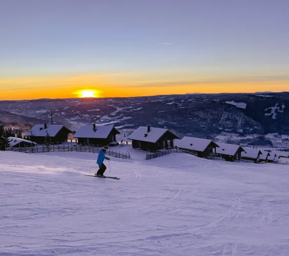 Ein Skifahrer fährt eine verschneite Piste hinab, vor Hütten und Bergen im warmen Licht des Sonnenuntergangs..