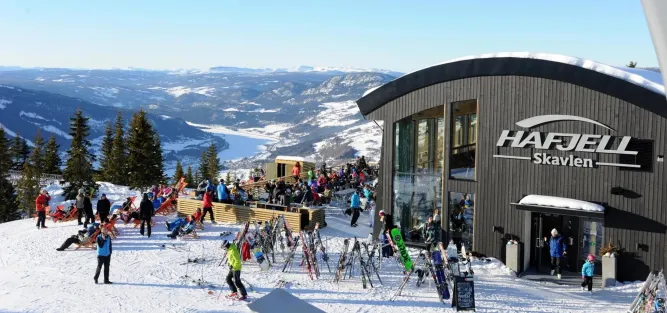 Skigebiet mit moderner Hütte, vielen Skifahrern, abgestellten Skiern und Blick über verschneite Berge und See..