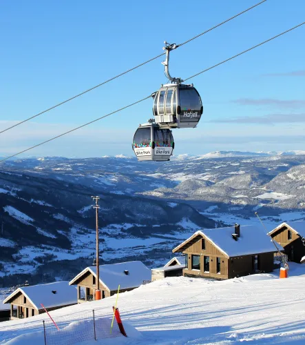 Zwei Gondeln einer Seilbahn schweben über einem verschneiten Skigebiet mit Hütten im Vordergrund und Bergen im Hintergrund..