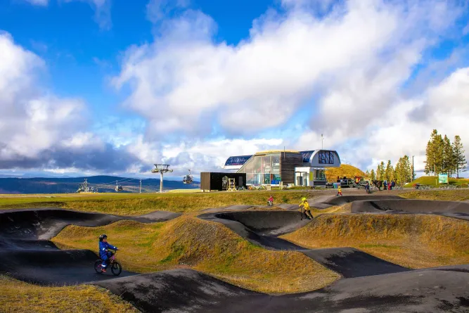 Kinder fahren auf einer Pumptrack-Strecke nahe einer Bergstation mit Gondelbahn unter blauem Himmel und Wolken..