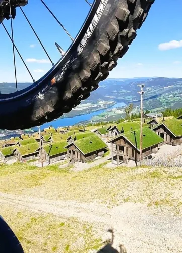 Nahaufnahme eines Mountainbike-Reifens vor grünen Berghütten, Tal und See unter blauem Himmel..