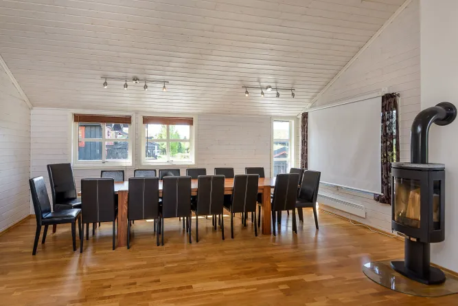 Lichte houten eetkamer met lange tafel, zwarte stoelen, drie ramen, een projectiescherm en een houtkachel..