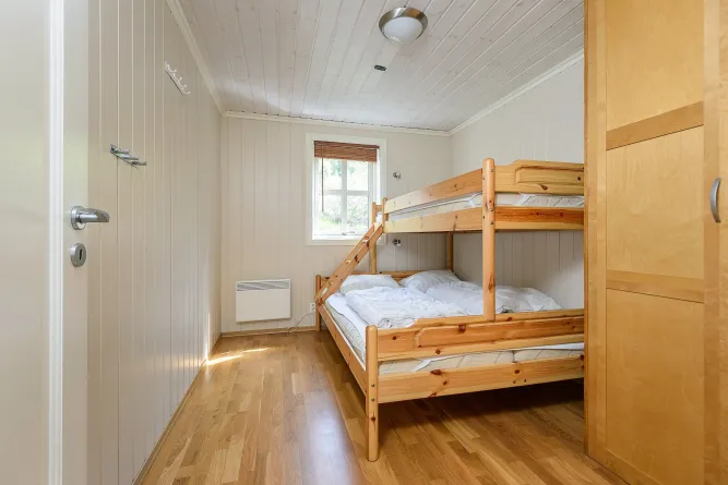 Kleine lichte slaapkamer met houten stapelbed, wit beddengoed, raam met rolgordijn en houten vloer..