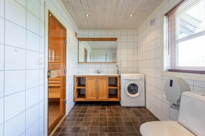 Heldere badkamer met witte tegels, houten wastafelmeubel, spiegel, wasmachine, toilet en sauna achter glazen deur..