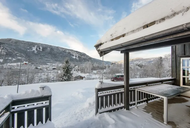 Besneeuwd houten terras met tafel en dakrand, uitkijkend over een winterdal met bergen onder blauwe lucht..