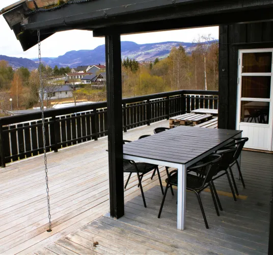 Houten terras met tafel en stoelen bij een donkere chalet, met uitzicht op dorp, bomen en bergen..