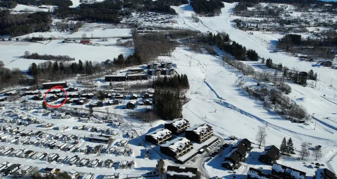 Luchtfoto van besneeuwd dorp met huizen en appartementen naast een brede skipiste; een rood omcirkeld gebouw links..