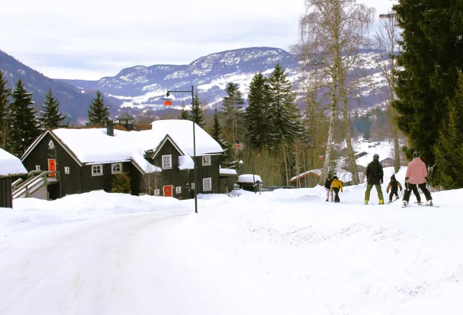 Besneeuwde dorpsweg met donker houten chalet, dennenbomen en berglandschap; snowboarders lopen omhoog..
