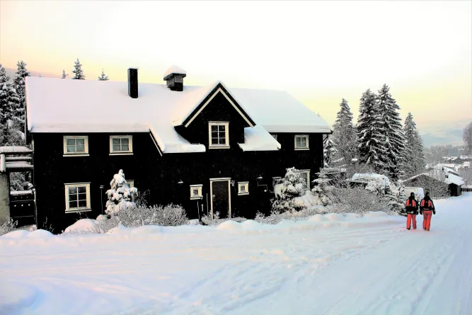 Zwart huis met besneeuwd dak naast een wit winterpad; twee wandelaars in rode kleding lopen langs sparrenbomen..