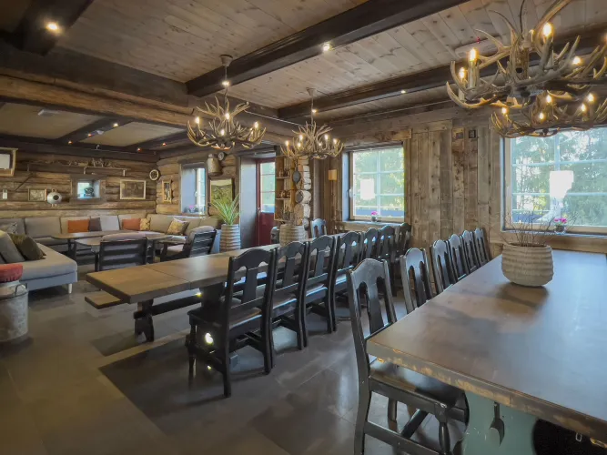 Rustieke houten eetzaal met lange tafels en stoelen, geweienlampen aan het plafond en ramen met daglicht..