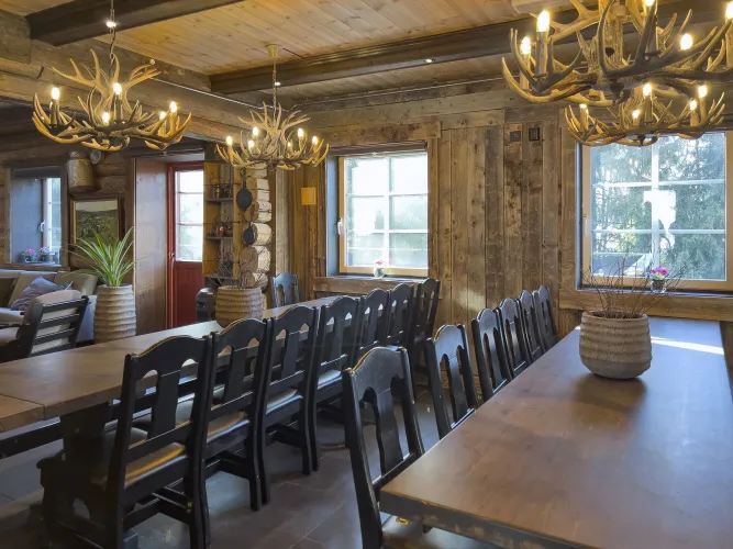 Interieur van houten eetkamer met lange tafels, zwarte stoelen, gewei-kroonluchters en ramen met daglicht..