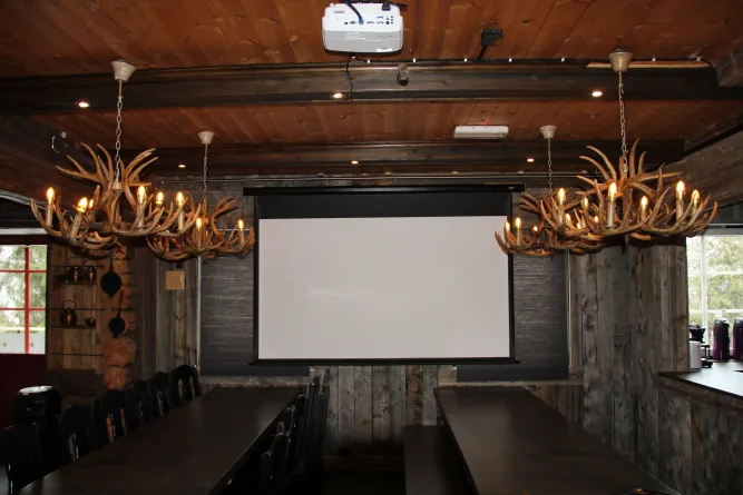 Rustieke houten ruimte met lange tafels, stoelen, geweikroonluchters en een projectiescherm aan de wand..