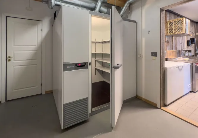 Witte inloopkoelcel met open deur en lege schappen in een werkruimte, met ventilatiebuizen en keukenhoek rechts..