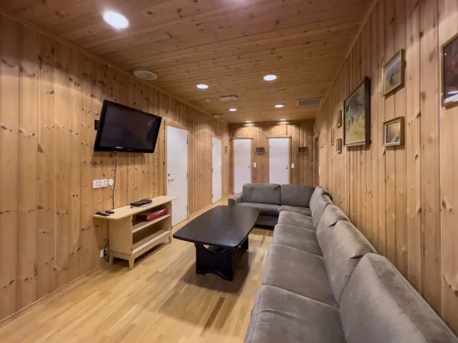 Gezellige lounge met houten wanden en plafond, grijze hoekbank, salontafel en tv aan de wand..