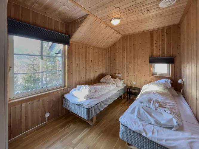 Houten slaapkamer met twee eenpersoonsbedden, rommelige witte dekens, nachtkastje en ramen met zicht op bomen..