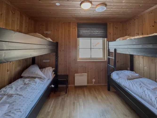 Kleine houten slaapkamer met twee stapelbedden, opgemaakt beddengoed, raam met rolgordijn en lampen aan het plafond..