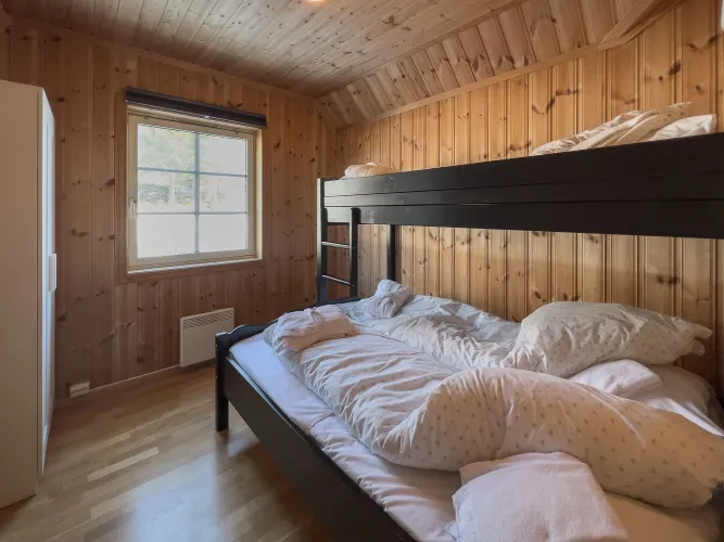 Houten slaapkamer met zwart stapelbed, rommelig beddengoed, raam met daglicht en witte kast links..