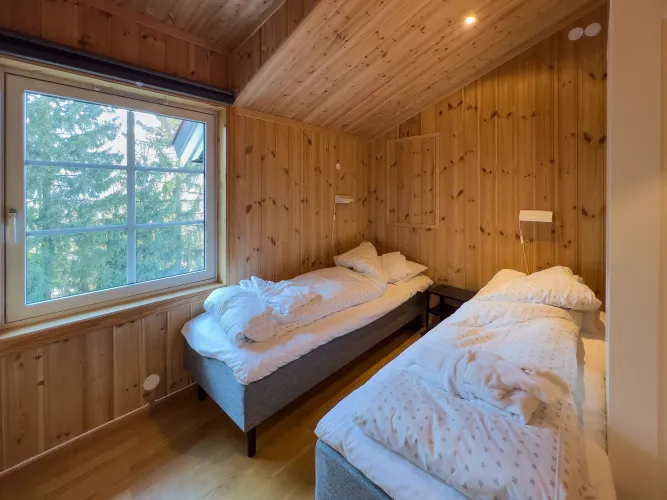 Houten slaapkamer met twee eenpersoonsbedden, wit beddengoed en een groot raam met uitzicht op bomen..