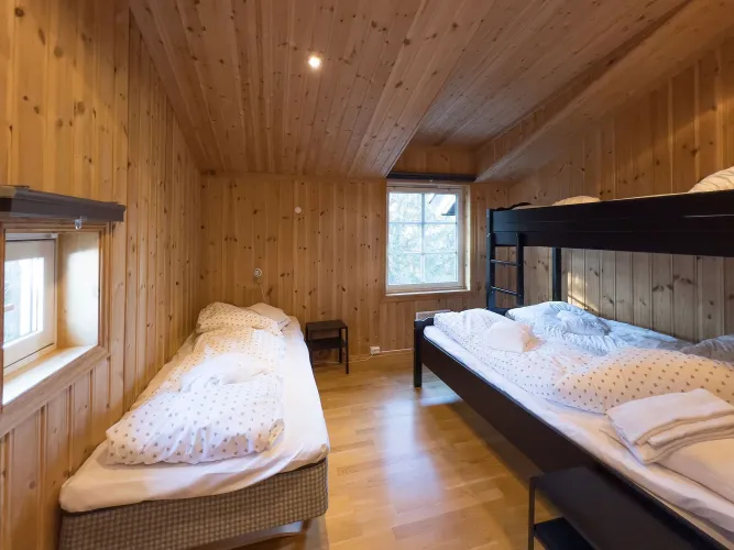 Gezellige houten slaapkamer met een eenpersoonsbed en een stapelbed, opgemaakte bedden en ramen met daglicht..