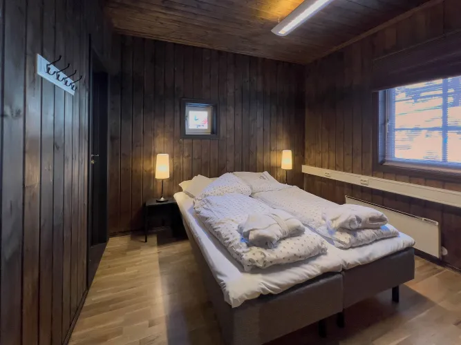 Houten slaapkamer met tweepersoonsbed, witte dekens en handdoeken, twee nachtlampjes en raam met jaloezieën..