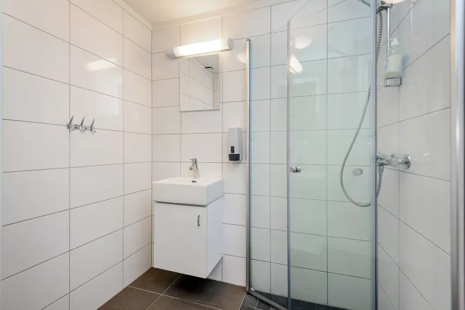 Witte betegelde badkamer met wastafelmeubel, spiegel en lamp, plus glazen douchewand en handdouche..