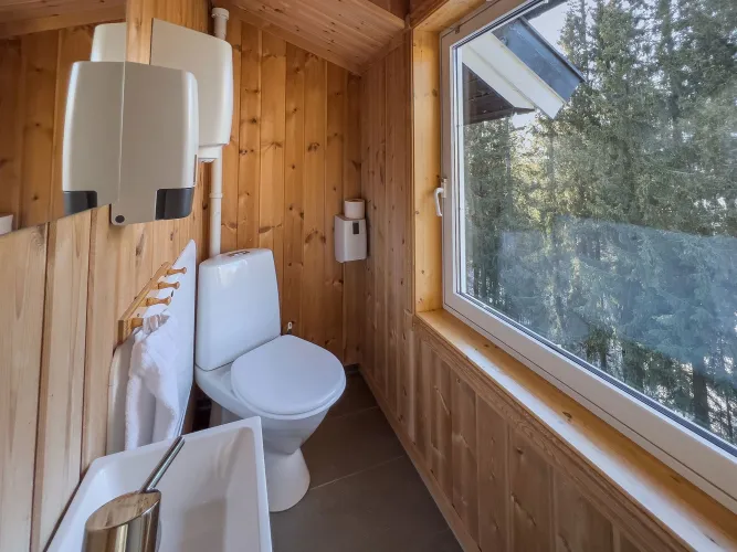 Klein houten toilet met wc, wastafel en dispensers, naast een groot raam met uitzicht op bomen buiten..