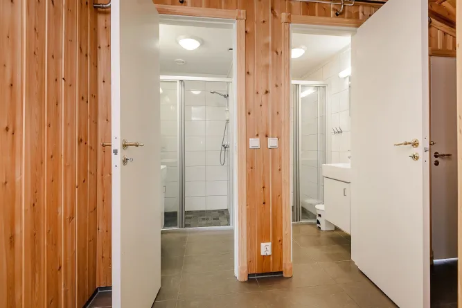 Houten gang met twee open deuren naar badkamers met douchecabines, witte tegels, wastafel en plafondlampen..