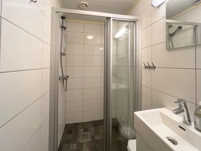 Witte betegelde badkamer met glazen douchecabine, douchekop, wastafel met kraan en spiegel aan de wand..