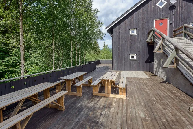 Groot houten terras met picknicktafels naast een donker houten gebouw met rode deur, omringd door bomen..