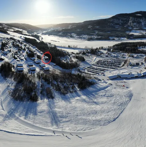 Luchtfoto van een besneeuwd dal met huizen, bosstroken, parkeerterrein en een brede skipiste in de zon..