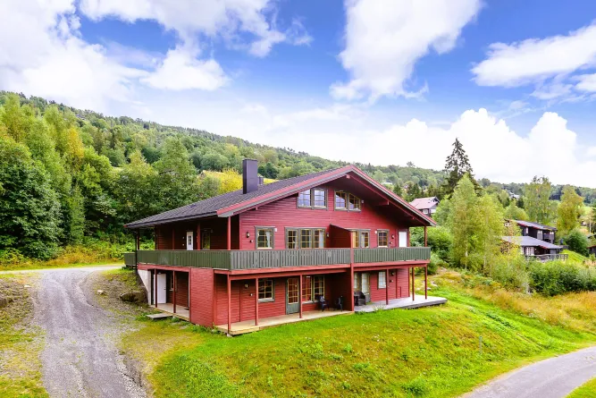 Ein rotes Holzhaus mit Balkon steht auf einer grünen Wiese, umgeben von Waldhängen unter blauem Himmel mit Wolken..