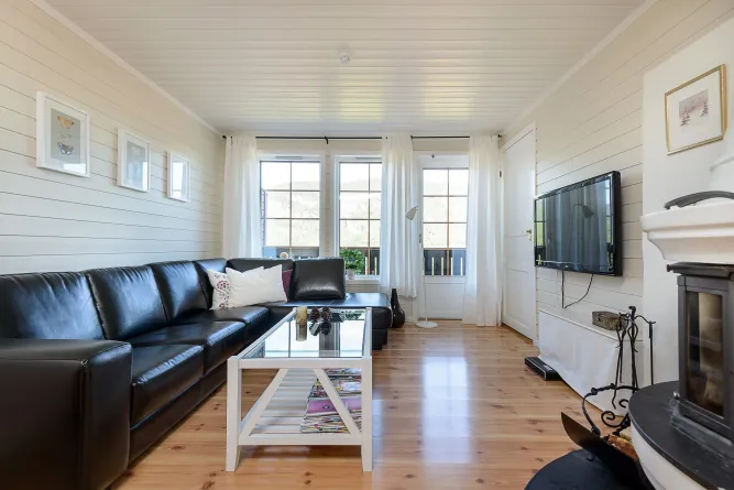 Helles Wohnzimmer mit schwarzer Ledercouch, Glastisch, Holzfußboden, weißen Vorhängen, TV an der Wand und Kamin..