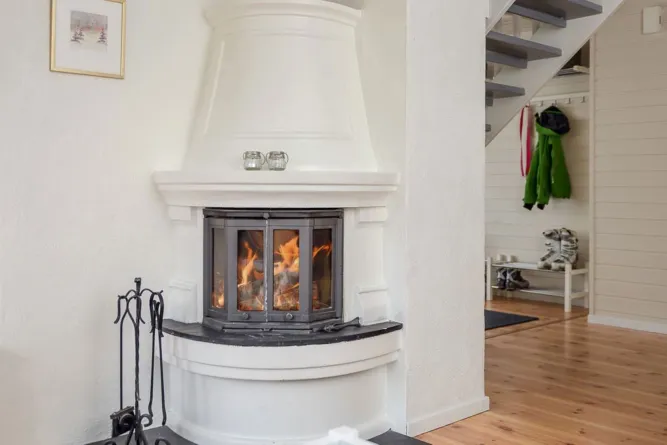 Weißer Kamin mit Feuer hinter Glasscheiben, daneben Kaminbesteck; rechts Flur mit Treppe, Jacken und Schuhen..