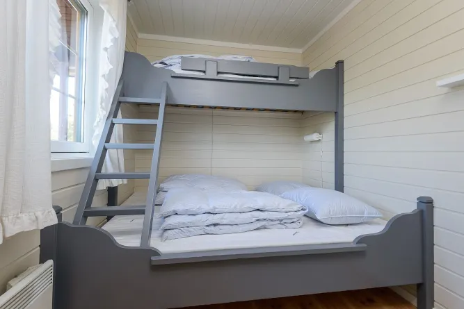 Schlafzimmer mit grauem Etagenbett und Leiter, frisch gestapelter Bettwäsche und Kissen, neben einem Fenster mit Vorhängen..