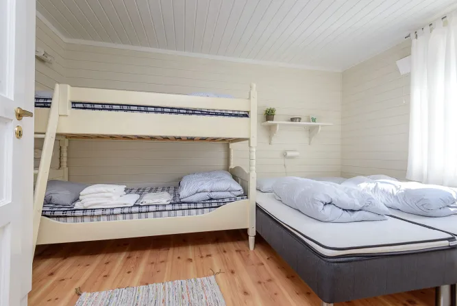 Helles Schlafzimmer mit weißem Etagenbett, Doppelbett, Holzfußboden, kleinem Regal und Vorhang am Fenster..