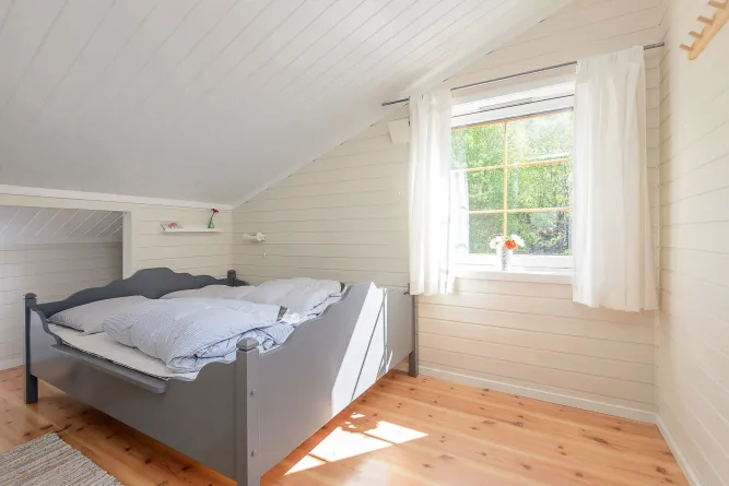 Helles Schlafzimmer im Dachgeschoss mit grauem Bett, Holzfußboden, weißer Vertäfelung und Fenster mit Vorhängen..