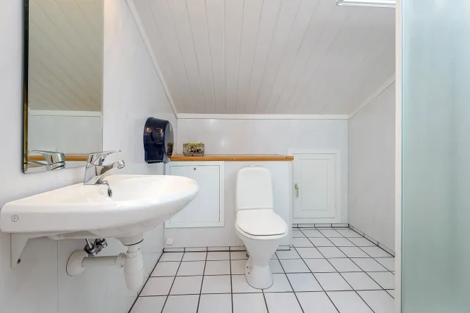 Weiß gefliestes Badezimmer mit Waschbecken links, Spiegel, WC mittig und schräger Decke; rechts Milchglaswand..