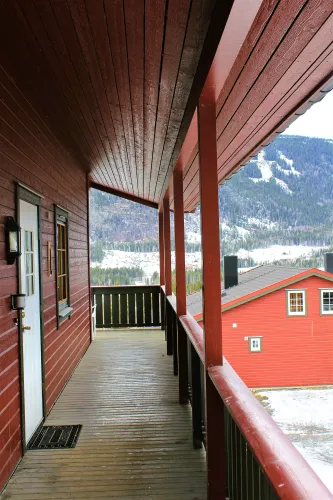 Überdachter Holzbalkon eines roten Hauses mit Geländer und Säulen, im Hintergrund verschneite Berge und Wald..
