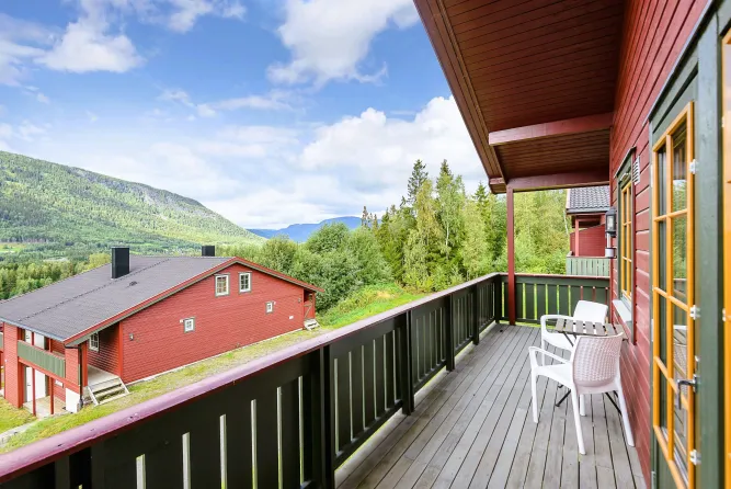 Holzbalkon mit weißen Stühlen neben roter Hütte, Blick auf grüne Berge, Wald und blauen Himmel mit Wolken..