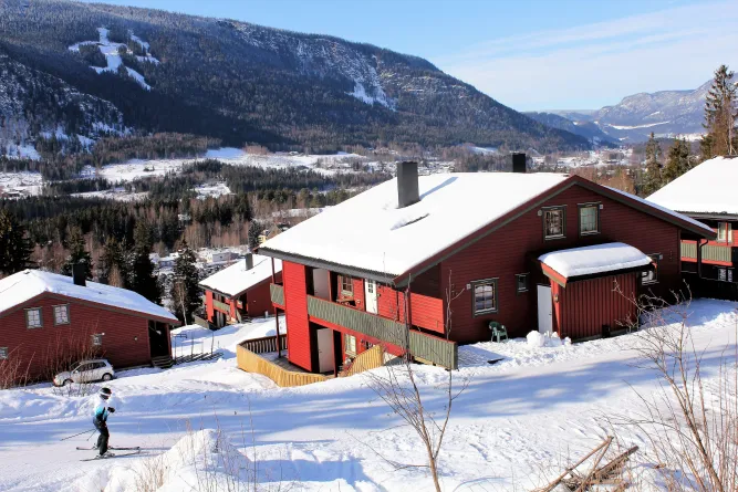 Rotes Holzhaus mit schneebedecktem Dach am Hang; im Hintergrund Wald, Tal und Berge unter blauem Himmel..