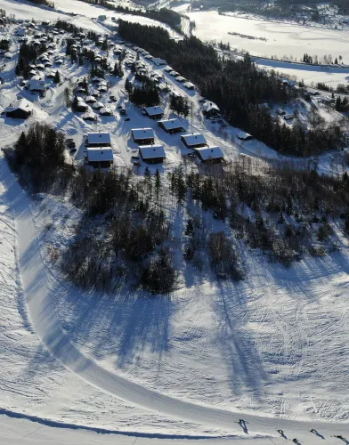 Luftaufnahme eines verschneiten Berghangs mit verstreuten Hütten, Waldstreifen und Spuren im Schnee bei Sonne..