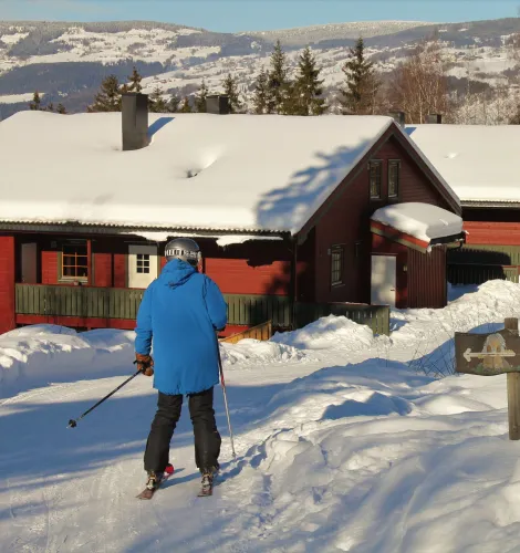 Eine Person mit Ski und blauem Anorak steht auf verschneitem Weg vor einer roten Hütte in Berglandschaft..