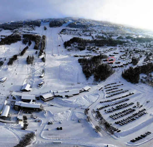 Luftaufnahme eines verschneiten Skigebiets mit Skipisten, Talstation, großem Parkplatz und verstreuten Chalets am Hang..